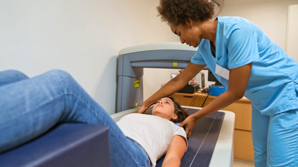 Female nurse preparing woman for bone density scan. Healthcare worker is looking at young patient lying on examination table. They are at hospital.