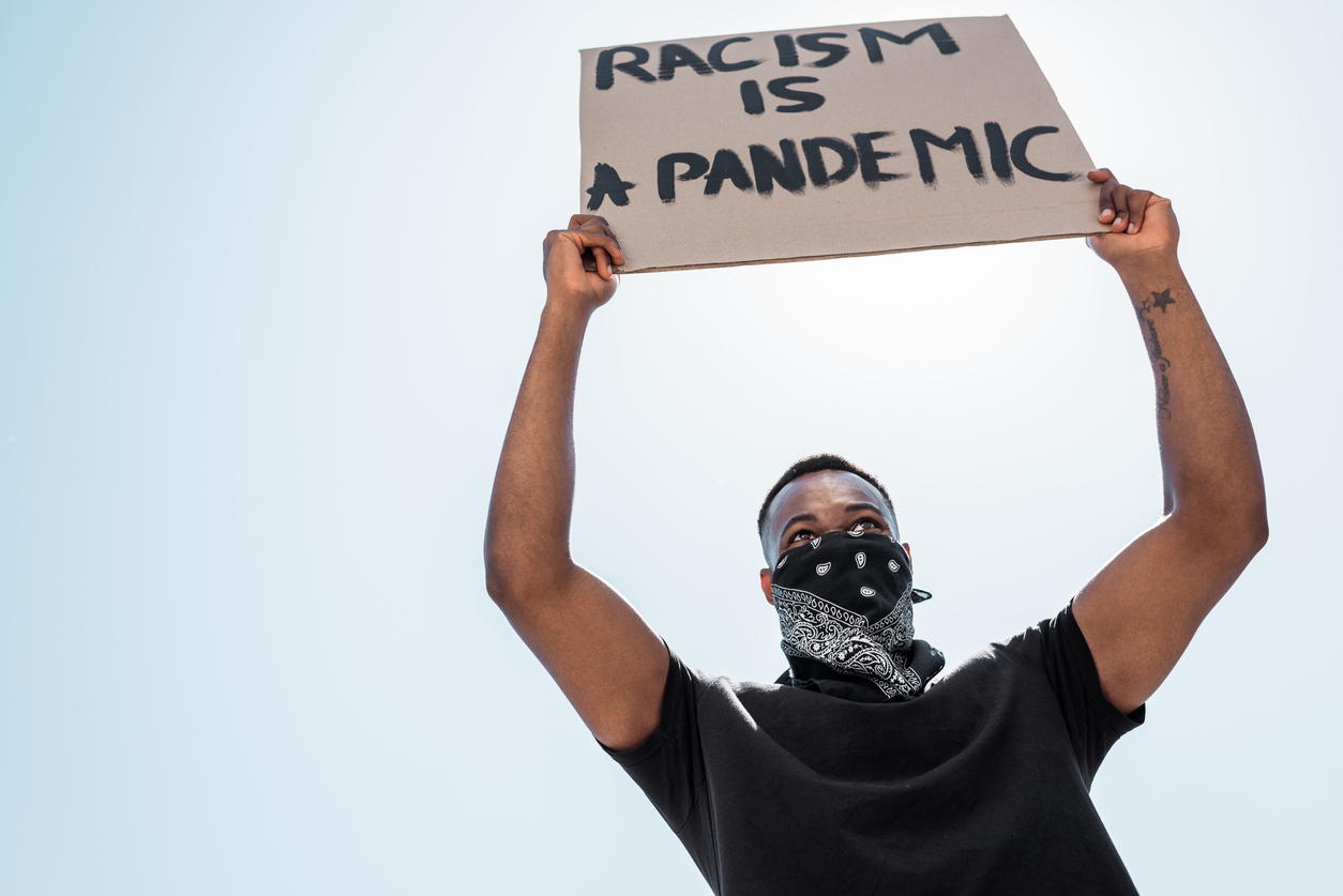 low angle view of african american man with scarf on face holding placard with racism is a pandemic lettering against blue sky