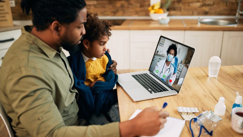 Black father and daughter communicating with a doctor via video call from home during COVID-19 pandemic. Focus is on girl.