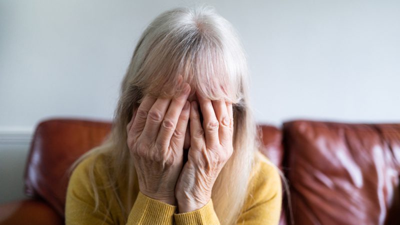 Color image depicting a sad, depressed-looking senior woman in her 70s, and of Caucasian ethnicity, sitting with her head in her hand at home.