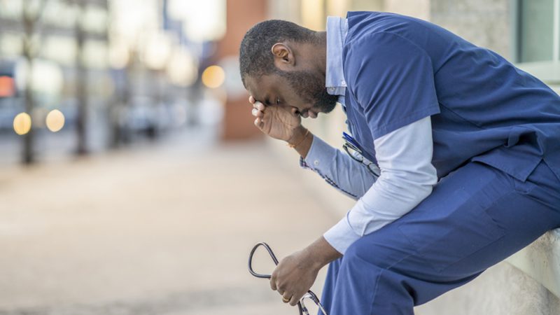 A male doctor sitting outside the hospital building trying to catch some rest. He is wearing blue medical scrubs and is resting his head on his hand while holding a stethoscope with his hands.