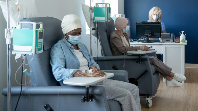 Two senior women sit side-by-side in their own chairs as they receive their cancer treatments intravenously. They are both dressed casually and have head scarves on as they sit looking across the room. A nurse can be seen working at a desk in the background.