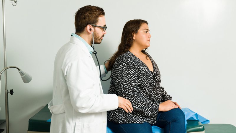 Caucasian doctor and latin female patient. Physician using the stethoscope to check the lungs of an ill woman